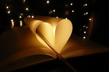 A book open on a table with the pages folded into the spine in the shape of a heart, with sparkling lights in the background.
